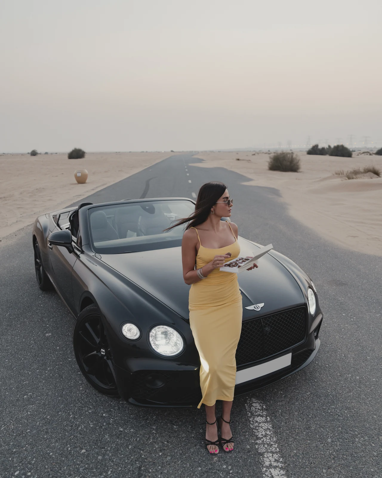 Yellow-dress model with the chocolate box in front of the convertible on a desert road.